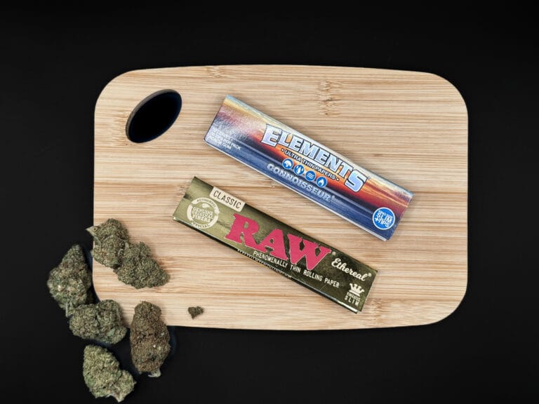 Häften av RAW Classic Ethereal och Elements Connoisseur på en skärbräda av bambu bredvid CBD-blommor